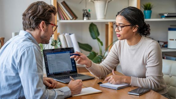 Crop faceless female freelancer in casual outfit sitting at table with cup of drink and surfing on laptop in light workspace
