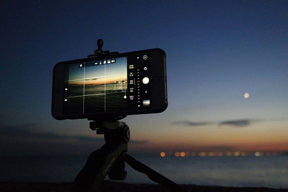 An evening scene of a smartphone on a tripod taking a photo of the sunset over the ocean.