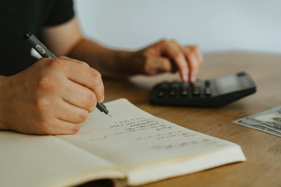 Close-up of hands writing calculations in a notebook with a calculator, focused on budgeting or financial work.