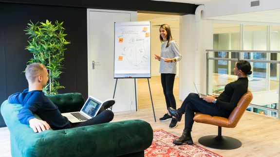 Three adults engaged in a casual office meeting with laptops and a flipchart.