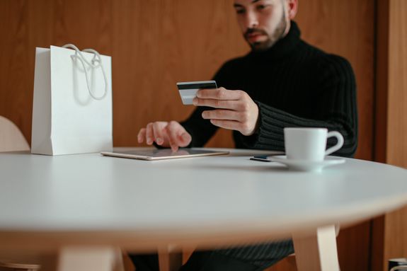 Crop focused bearded young man using tablet while doing online purchase with credit card sitting at table in modern cafe with coffee cup and shopping bag