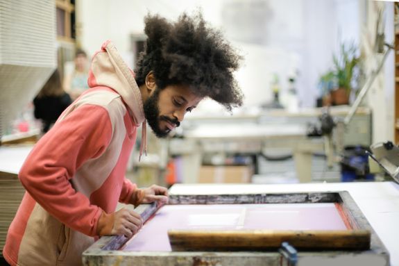 Side view of creative ethnic male in casual hoodie standing near workbench and working with wooden frame in creative workshop