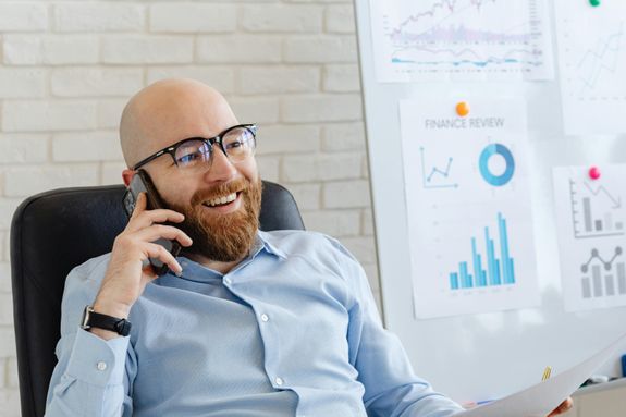 Bald businessman in glasses talks on phone, smiling at office with charts.