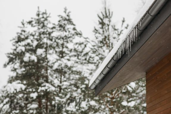 Stunning winter scene of icicles hanging from a snow-covered roof with snowy pines in the background.