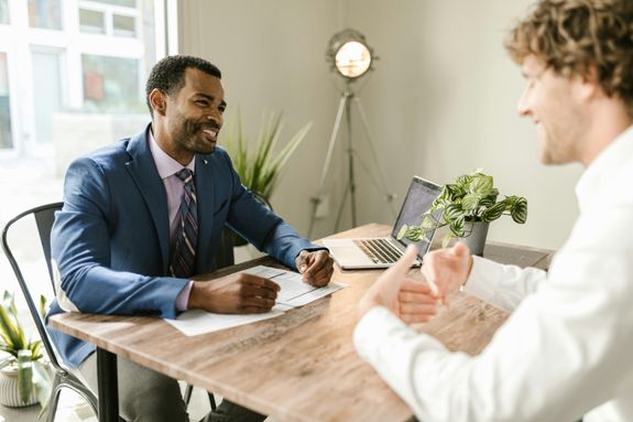 Two businessmen engaged in a positive conversation at an office desk with a laptop.