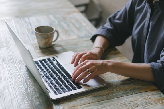 Crop faceless female freelancer in casual outfit sitting at table with cup of drink and surfing on laptop in light workspace