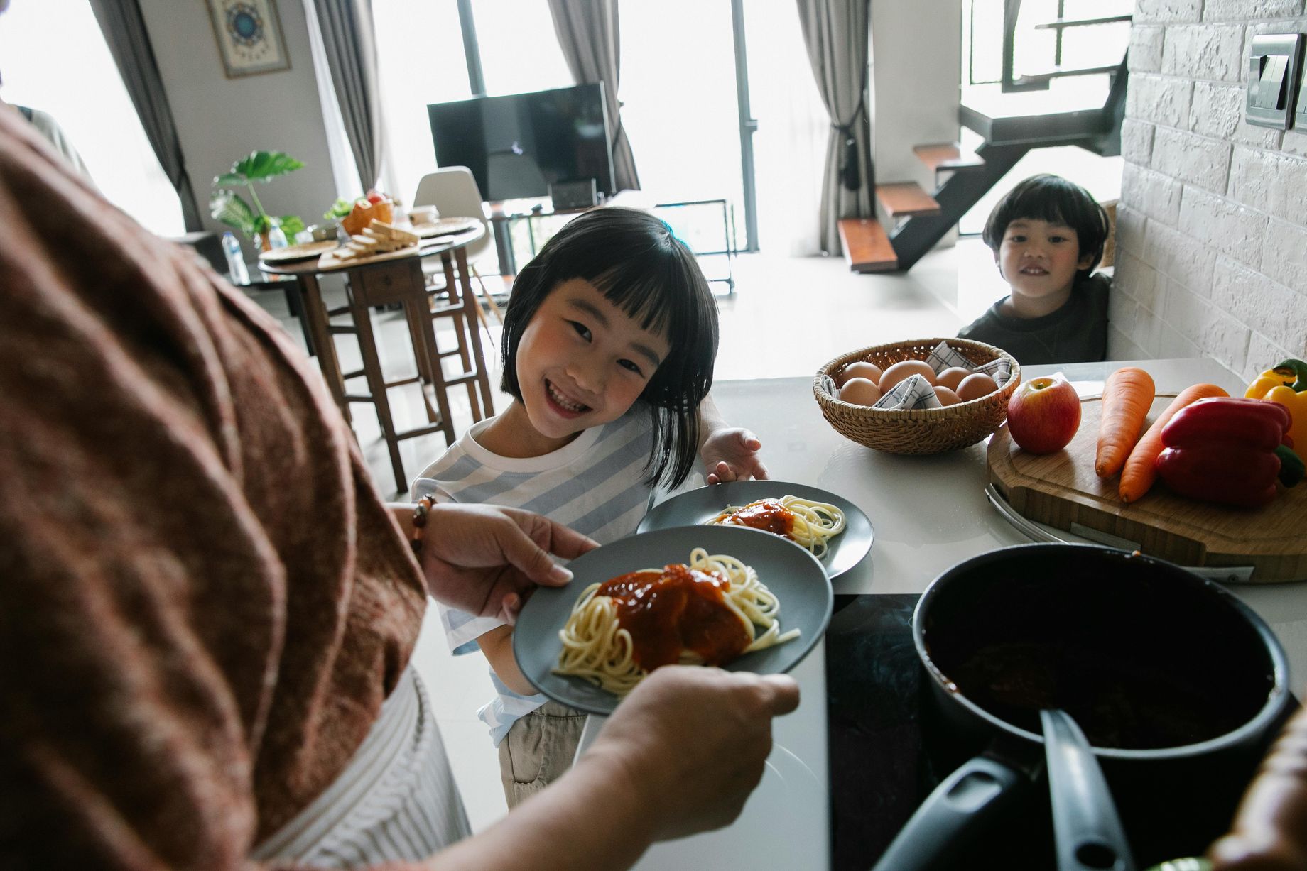 High angle of hungry cute little Asian children smiling while waiting for lunch prepared by unrecognizable grandmother standing in kitchen