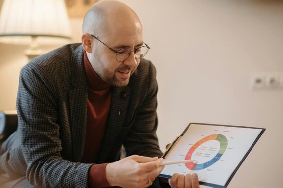 A man explains data using a color-coded chart on a clipboard in a cozy indoor setting.