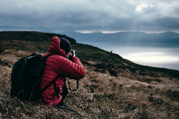 A hiker in a red jacket photographs a scenic mountain sunset by the sea.