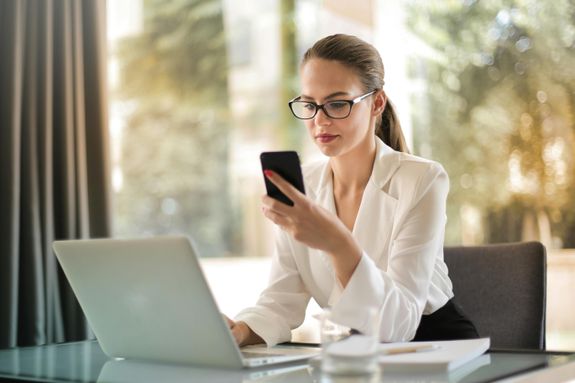 Professional woman using smartphone while working on laptop in modern office.