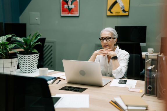 An elderly businesswoman in an office setting, smiling while using a laptop.