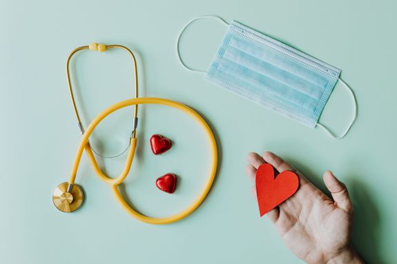 Top view of crop anonymous person hand with red paper heart on table with stethoscope and medical mask for coronavirus prevention