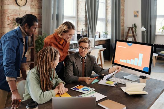 A Group of People Discussing Beside a Desktop with Graph on Screen