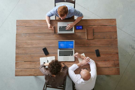 A team of three professionals collaborating at a wooden table with laptops and smartphones in a modern office.