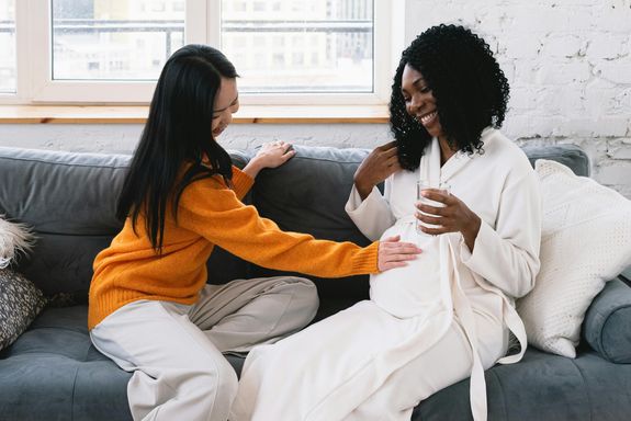 Cheerful Asian lesbian touching tummy of happy pregnant African American female while sitting on couch in living room at home