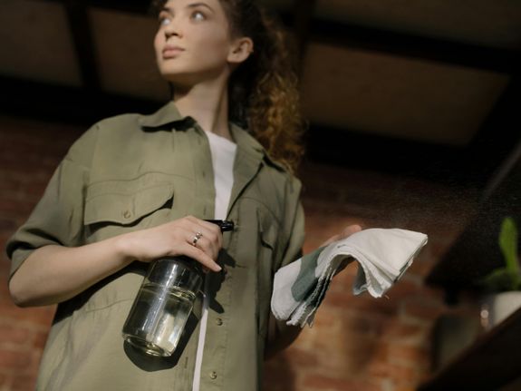 A woman with curly hair holds a cleaning spray and cloth in a cozy home setting.