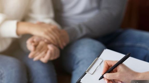 Crop unrecognizable psychotherapist taking notes in journal while interacting with woman on chair during psychological counseling indoors