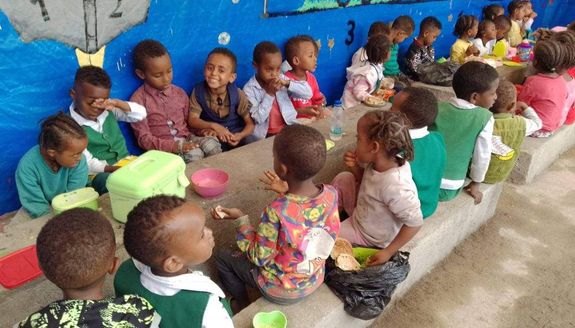 A group of school-age Ethiopian children eating lunch in front of a blue wall